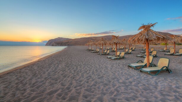 A beach with rows of sun loungers and thatched umbrellas, a calm sea, and a sunset in the background. - Aeolian Village Beach Resort (Lesvos)