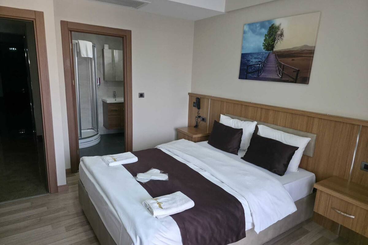 Ein Hotelzimmer mit einem großen Bett, einem hölzernen Kopfteil, einem Landschaftsbild, einem Badezimmer mit Glasdusche und einem Holzboden.
