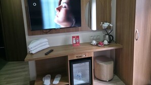 A hotel room with a wooden desk, a television mounted above it, a mirror, a small refrigerator, and a cup of coffee. - Hotel La SERA (Istanbul)