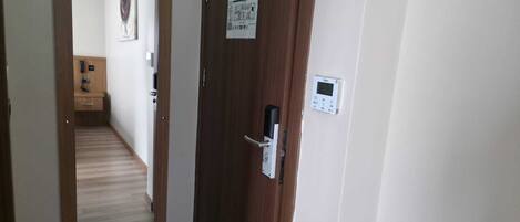 A hallway with a wooden door, a digital thermostat, and a mirror reflecting a bathroom with a toilet and a sink.