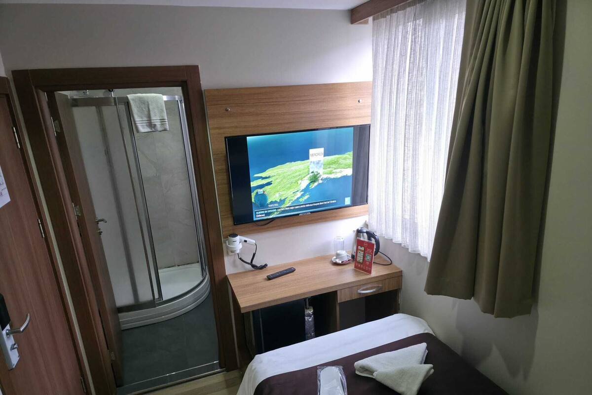 Ein Hotelzimmer mit Bett, Wandfernseher, Schreibtisch mit Fernbedienung, Telefon und einem Glas Wasser.