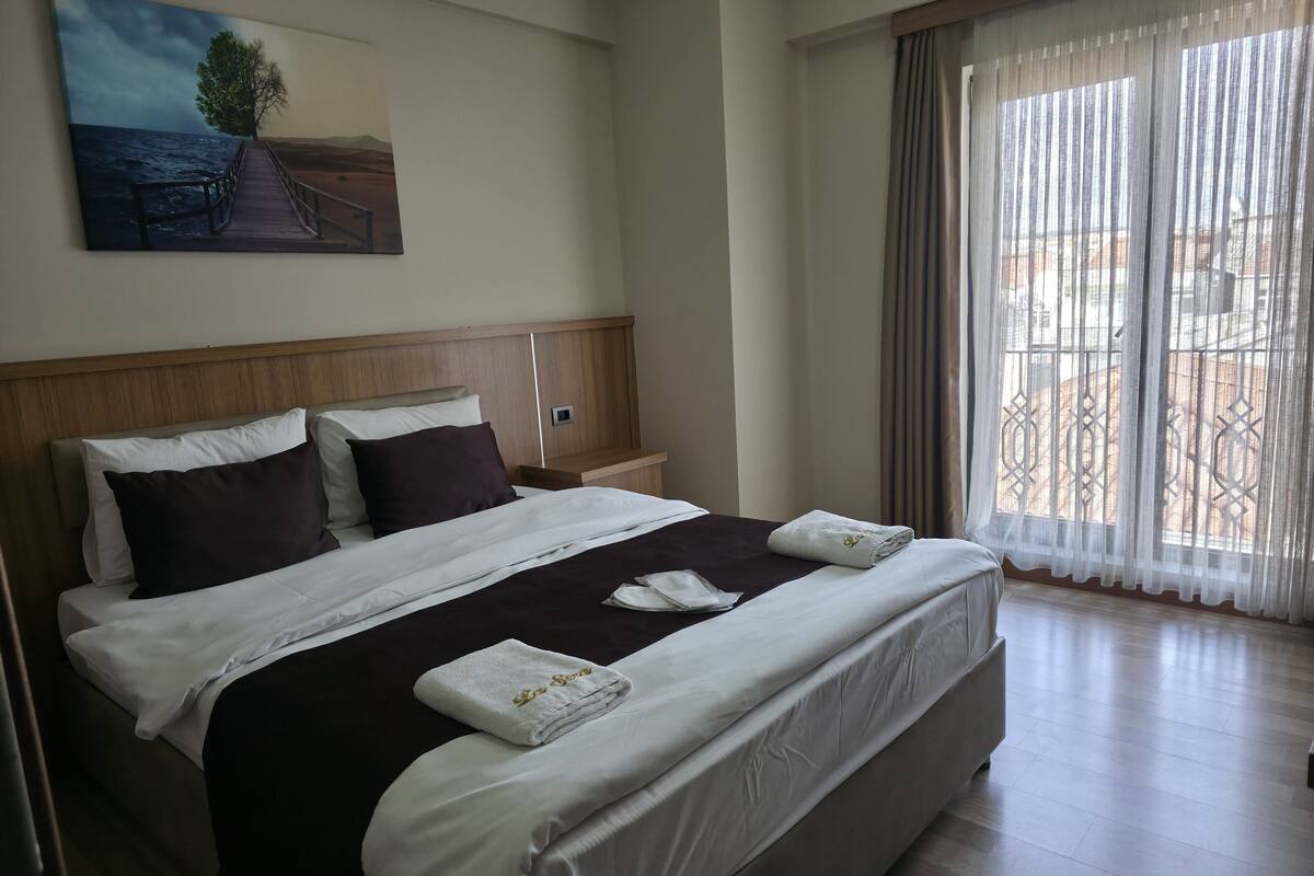 Ein Hotelzimmer mit einem großen Bett, einem Kopfteil, einem Nachttisch, einem Bild an der Wand und einem Fenster mit Vorhängen.