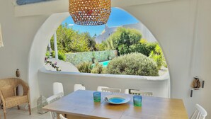 Zona de menjador amb taula de fusta i cadires blanques, una cadira de vimet i un llum de llanternatge, amb vista a un jardí i piscina.