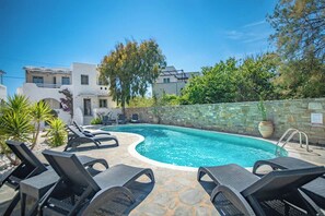A swimming pool with lounge chairs, a stone wall, and a white building with balconies. - A Perfect Home In Naxos (Stelida)