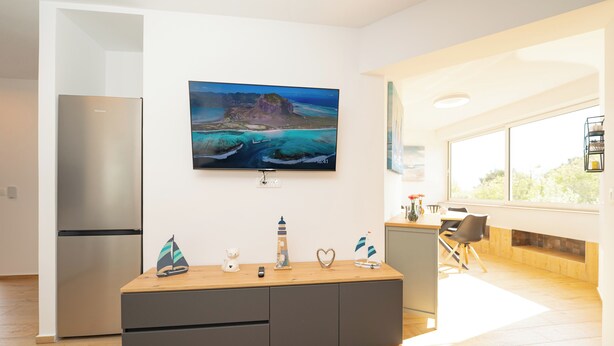 A modern kitchen with a built-in refrigerator, a wooden dining table, and a large window providing a view of the outdoors. - Apartments Adela I (Novalja)