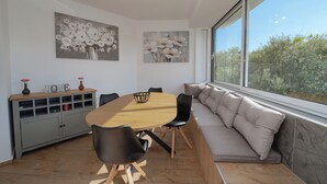 A modern dining area with a round wooden table, black chairs, and a grey cabinet with shelves. There are two wall-mounted paintings of flowers and a window with a view of greenery. - Apartments Adela I (Novalja)