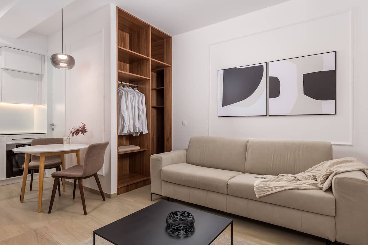 A modern living room with a beige sofa, a dining table with chairs, a wooden wardrobe, and abstract wall art.