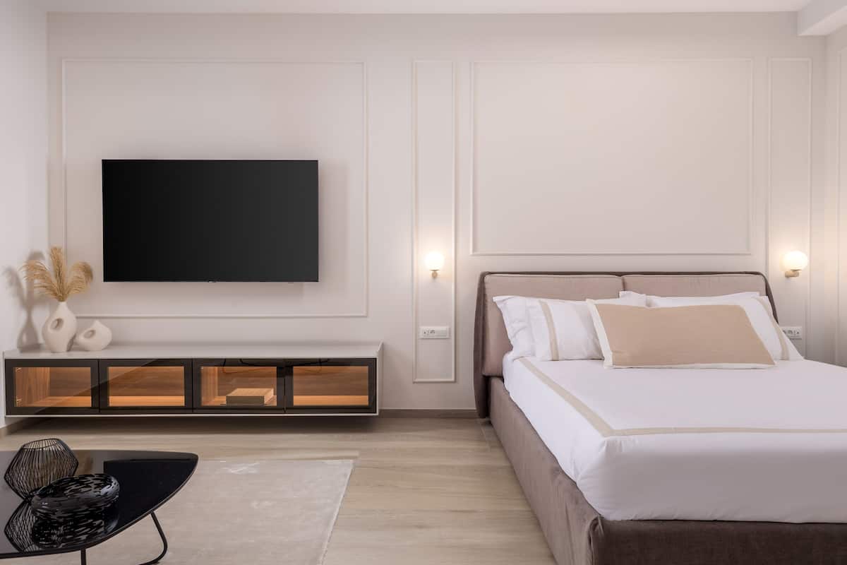 A modern bedroom with a bed, a flat-screen TV mounted on the wall, a bedside table with a lamp, and a small seating area with a chair and a table.