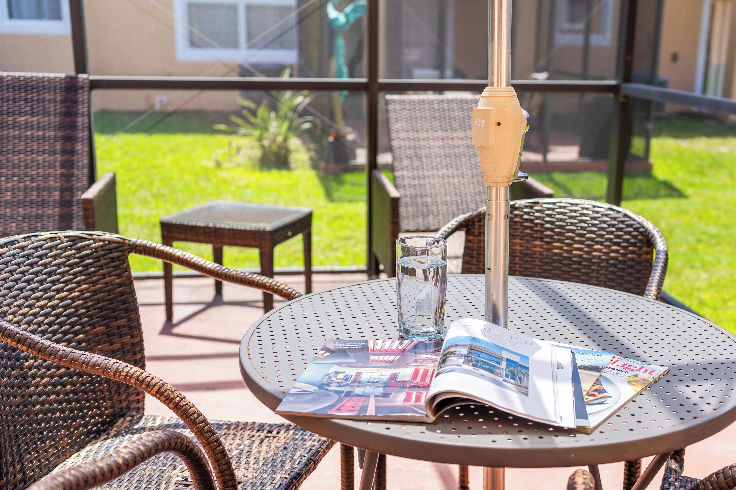 A round metal table with a magazine, a glass of water, and wicker chairs in a patio area with a view of a green lawn and a building in the background.