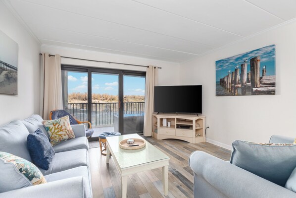 A modern living room with a sofa, coffee table, and TV stand. A large window offers a view of a wooden deck and beach.