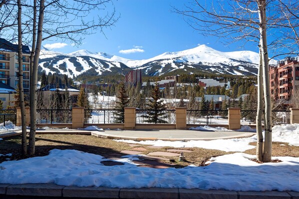 A snow-covered landscape with a mountain in the background, a paved area with trees, and buildings with balconies. - Hyatt 2BR Downtown | Steps to Peak 9 & Main St (Breckenridge)
