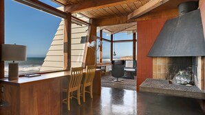 Un intérieur en bois avec une grande fenêtre donnant sur l’océan, une cheminée et un coin salon avec des fauteuils.