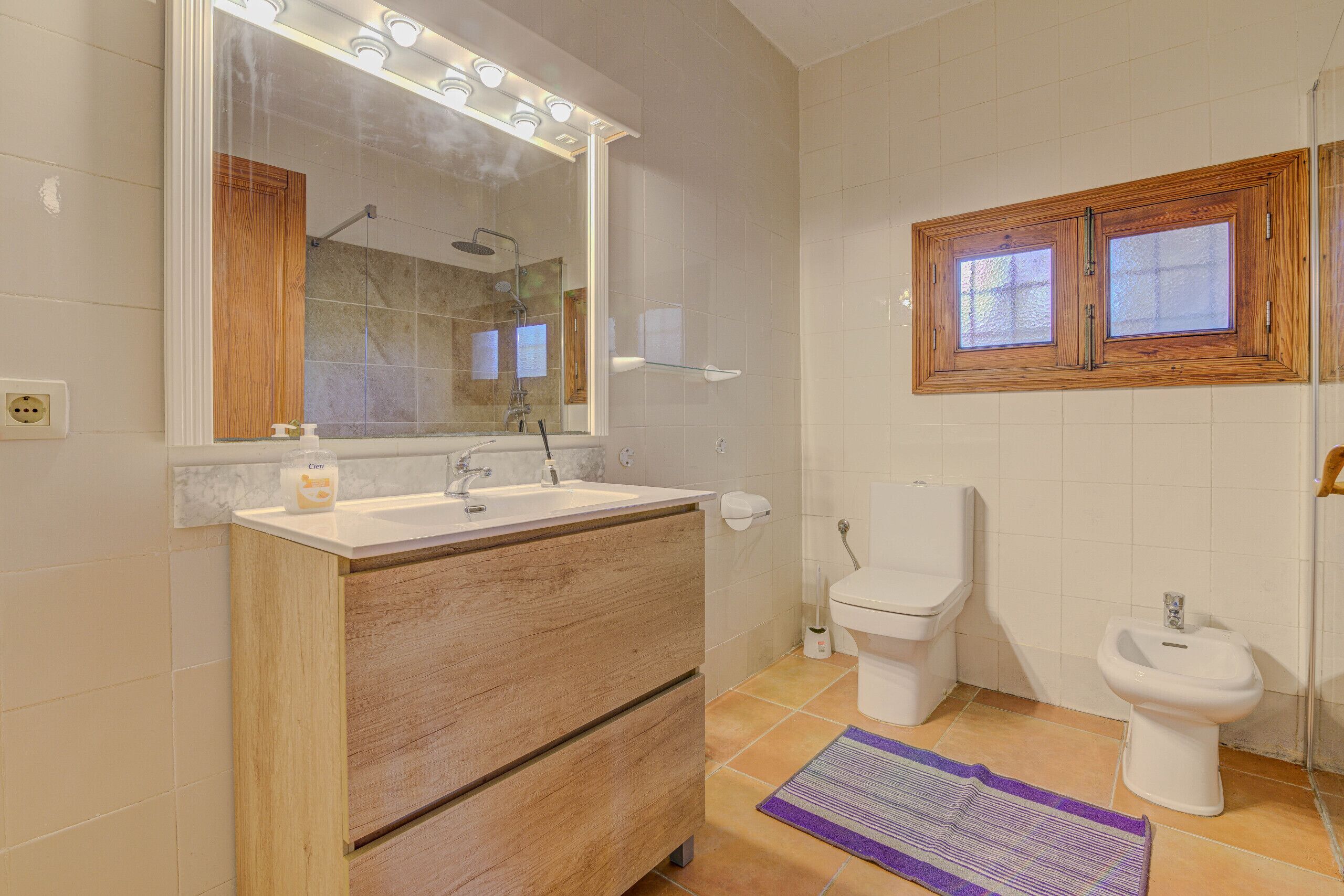A bathroom with a wooden vanity, a large mirror with lights, a toilet, a bidet, and a window with wooden frames.