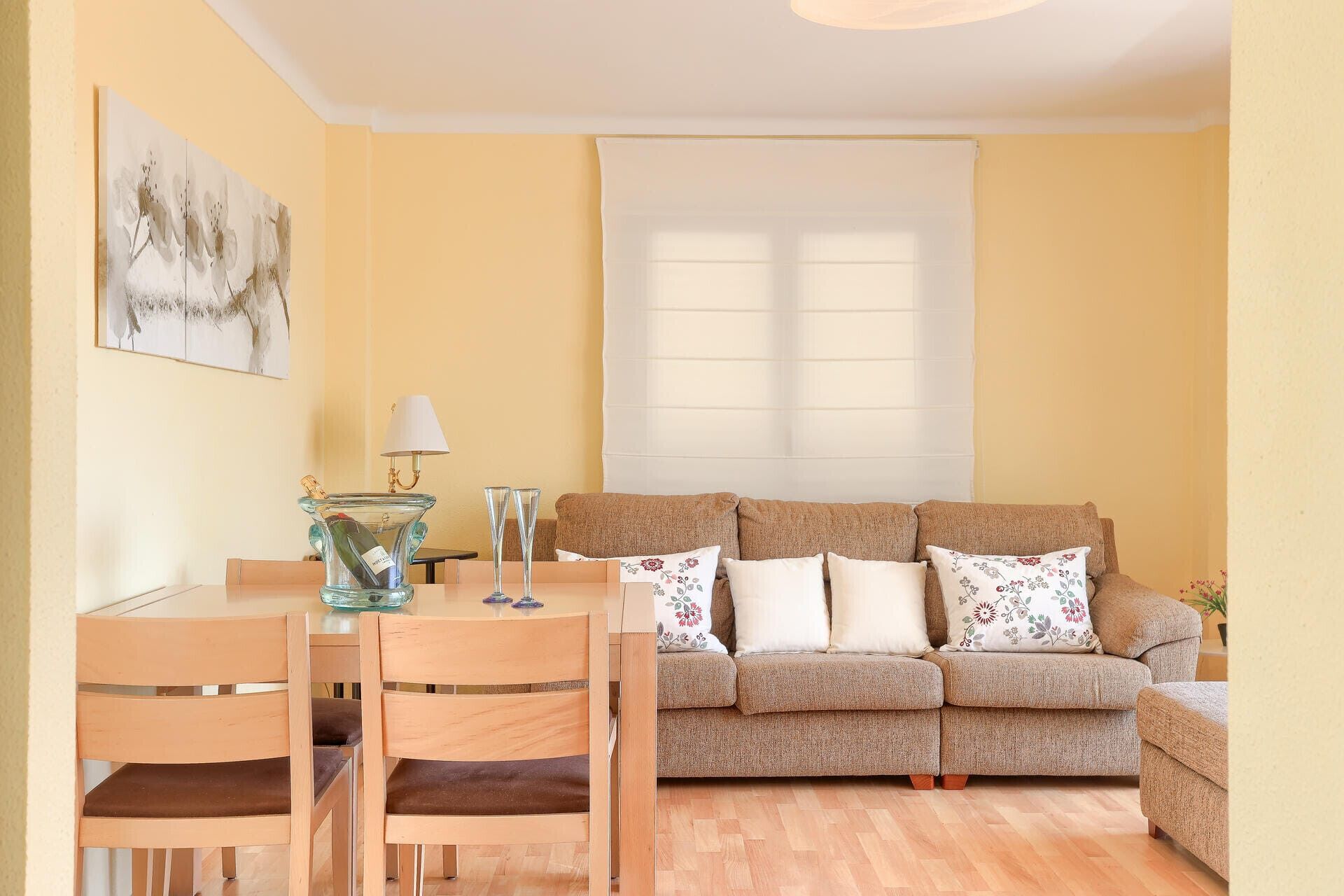 Een woonkamer met een beige bank, een eettafel met stoelen, een glazen karaf en twee glaasjes.