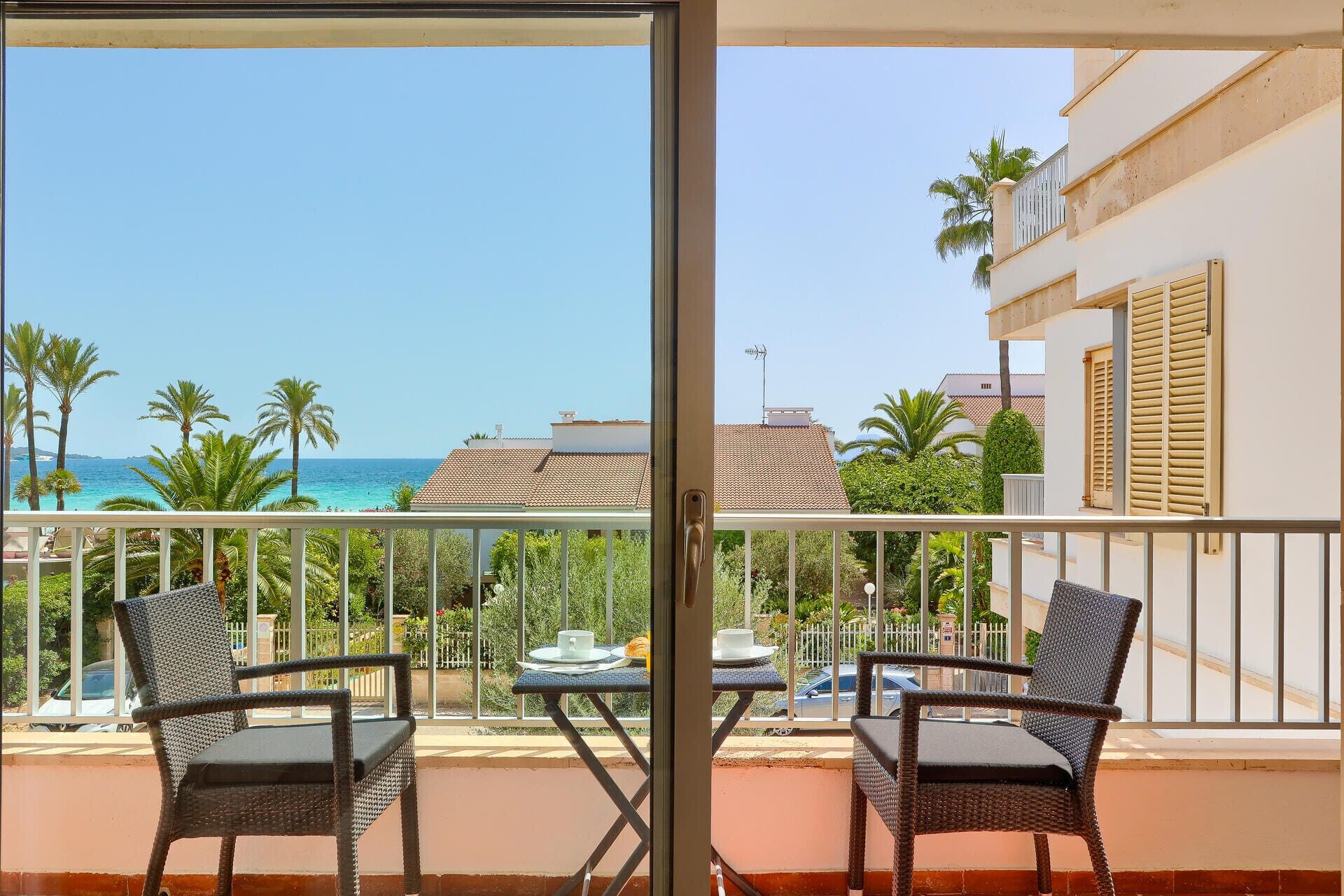Un balcon avec des fauteuils en osier et une table, donnant sur une plage bordée de palmiers et offrant une vue sur un ciel dégagé.