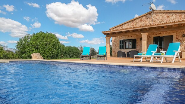 Una piscina con agua azul, rodeada por un edificio de piedra con techo de tejas, y dos sillas reclinables azules en el patio.