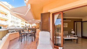 A balcony with a dining table and chairs, a lounge chair, and a sliding glass door leading to an interior room with a sofa and a table.