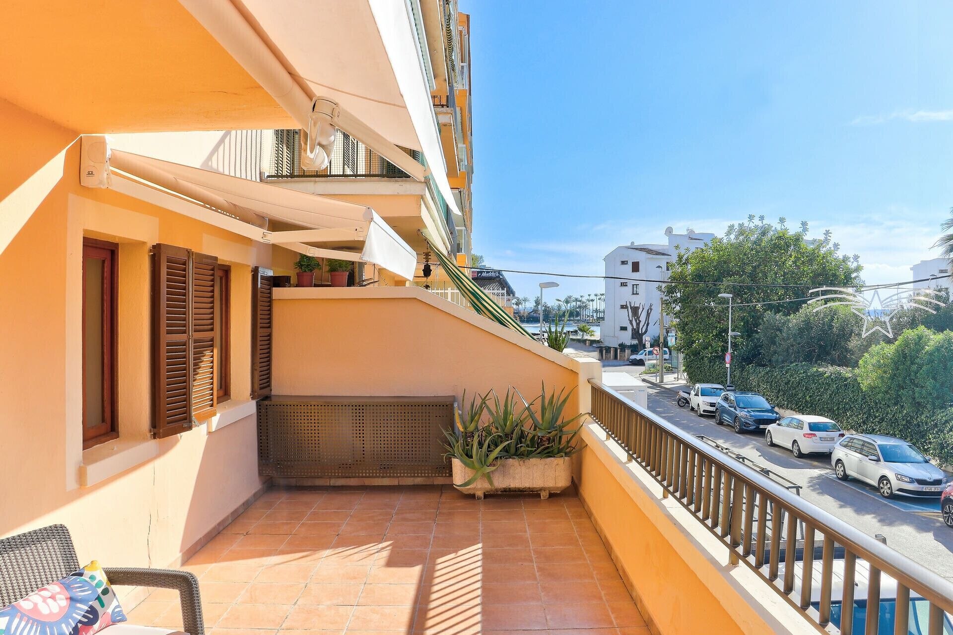 A balcony with a tiled floor, a chair, a potted plant, and a view of a street with parked cars and buildings.