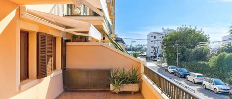A balcony with a tiled floor, a chair, a potted plant, and a view of a street with parked cars and buildings.