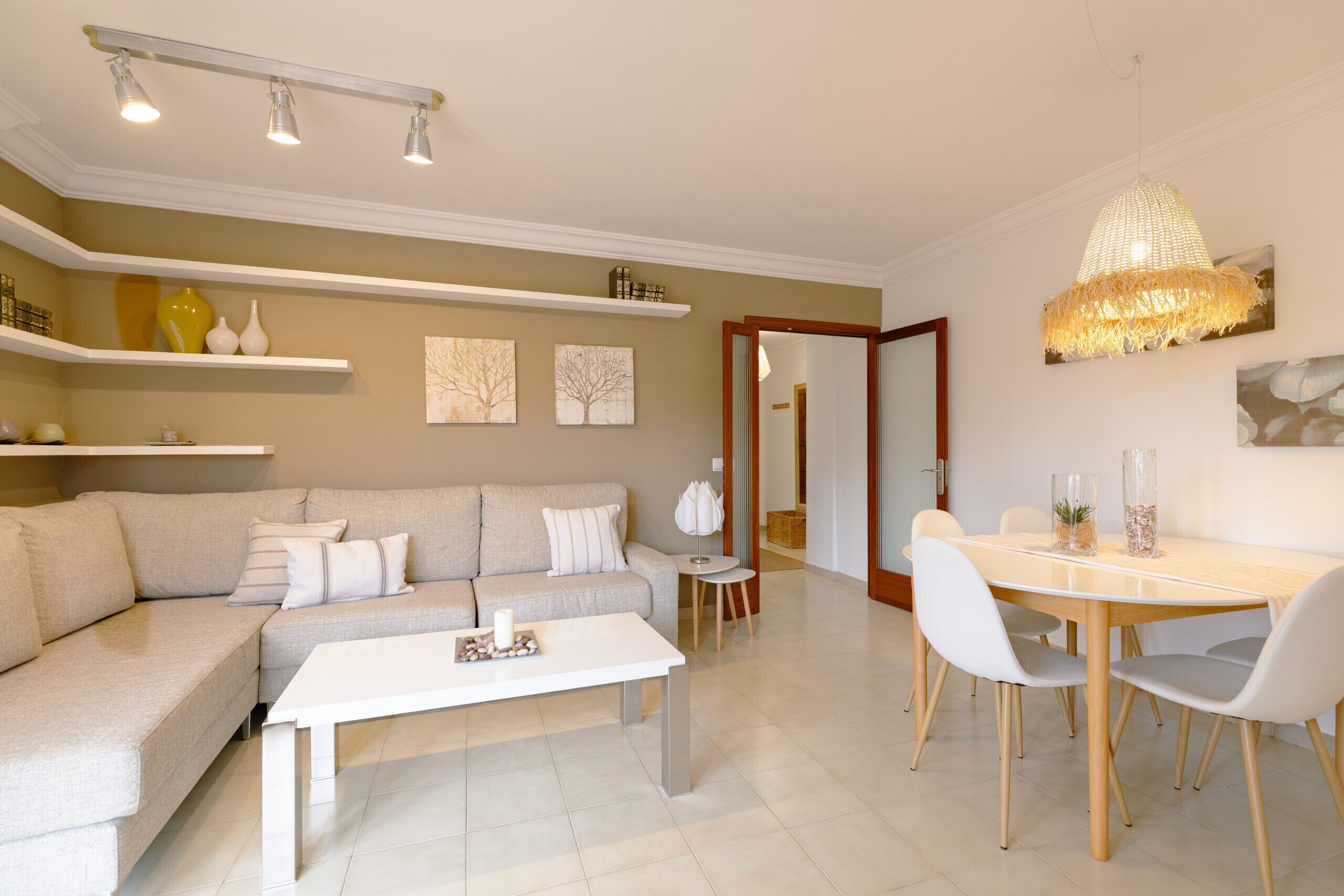 A living room with a sofa, coffee table, dining table, and chairs. There are shelves with decorative items and a hanging light fixture.