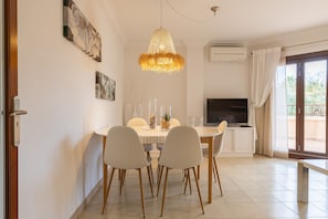 A dining area with a round table, white chairs, a chandelier, and a TV in the background.