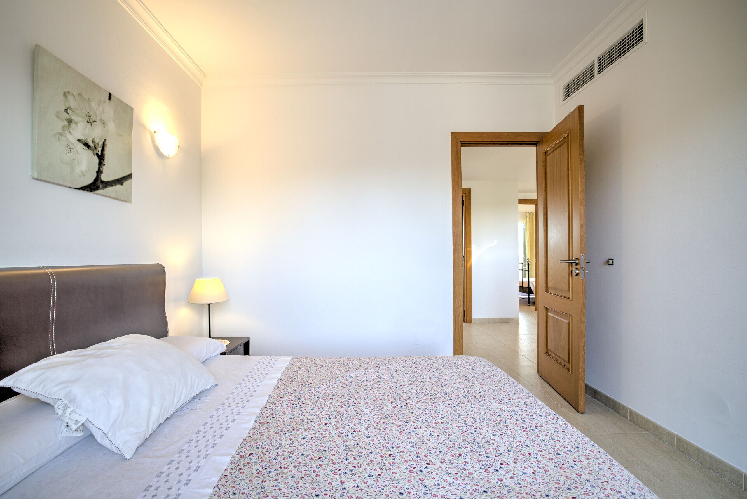 A hotel room with a bed, a nightstand, a lamp, a wall-mounted picture, and a door leading to another room.