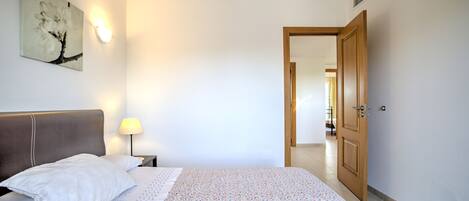 A hotel room with a bed, a nightstand, a lamp, a wall-mounted picture, and a door leading to another room.
