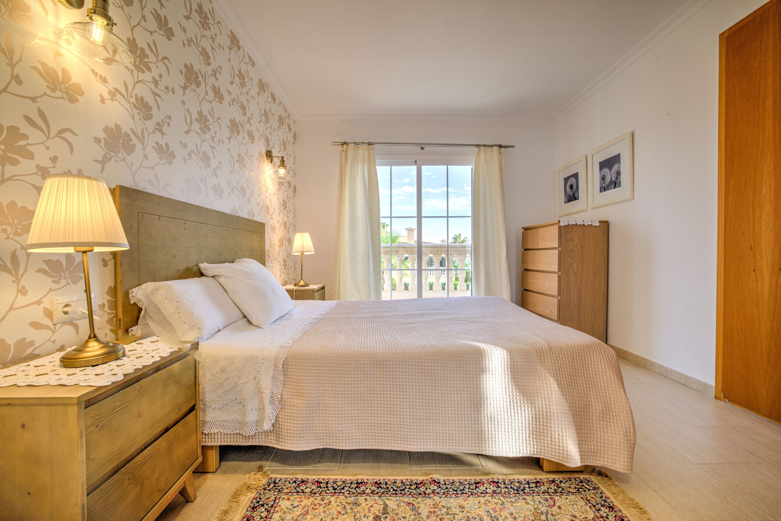 Een slaapkamer met een groot bed, twee bedlampen, een houten nachtkastje, een kantklosje, een vloerkleed met bloemenprint en een raam met gordijnen.