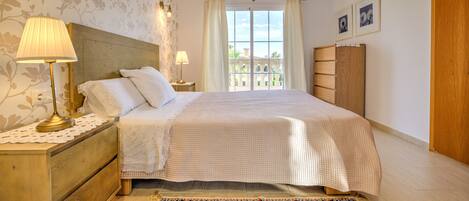 Een slaapkamer met een groot bed, twee bedlampen, een houten nachtkastje, een kantklosje, een vloerkleed met bloemenprint en een raam met gordijnen.