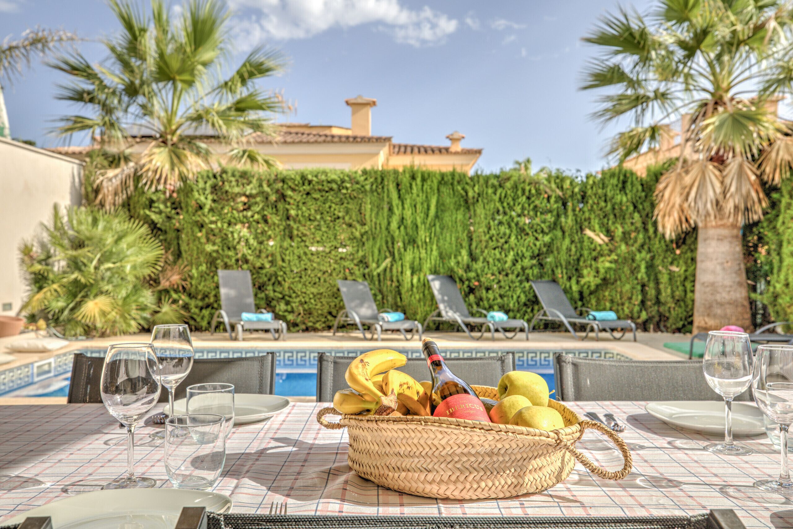 Une table dressée, sur laquelle se trouve un panier en osier contenant des fruits et une bouteille, donnant sur un espace piscine avec des chaises longues et une haie.