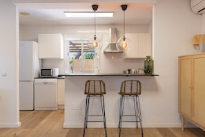 Una cocina moderna con armarios blancos, un microondas y un lavavajillas. Hay dos taburetes de bar con asientos de mimbre y estructura metálica negra.
