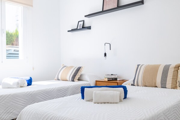 A hotel room with two beds, a bedside table with a lamp, a shelf with a picture, and a window with a view of greenery. - Orquidia by Rentallorca (Puerto de Alcudia)