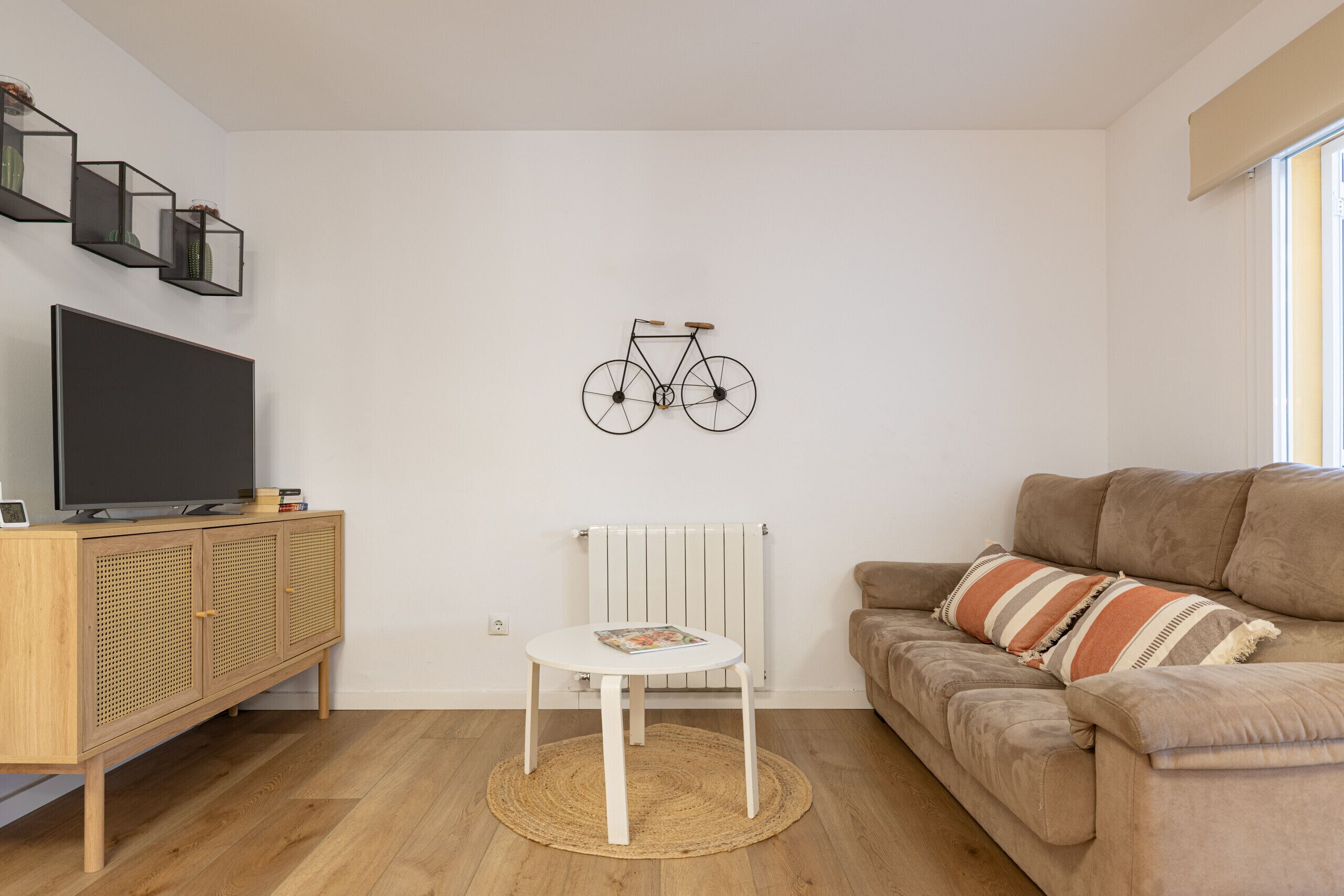 Uma sala de estar com televisão, um armário de madeira, uma mesa redonda, um sofá bege e uma bicicleta decorativa fixada na parede.