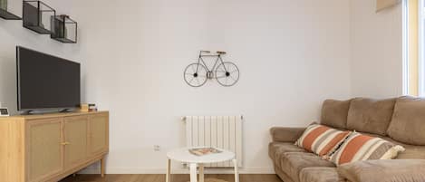 Uma sala de estar com televisão, um armário de madeira, uma mesa redonda, um sofá bege e uma bicicleta decorativa fixada na parede.