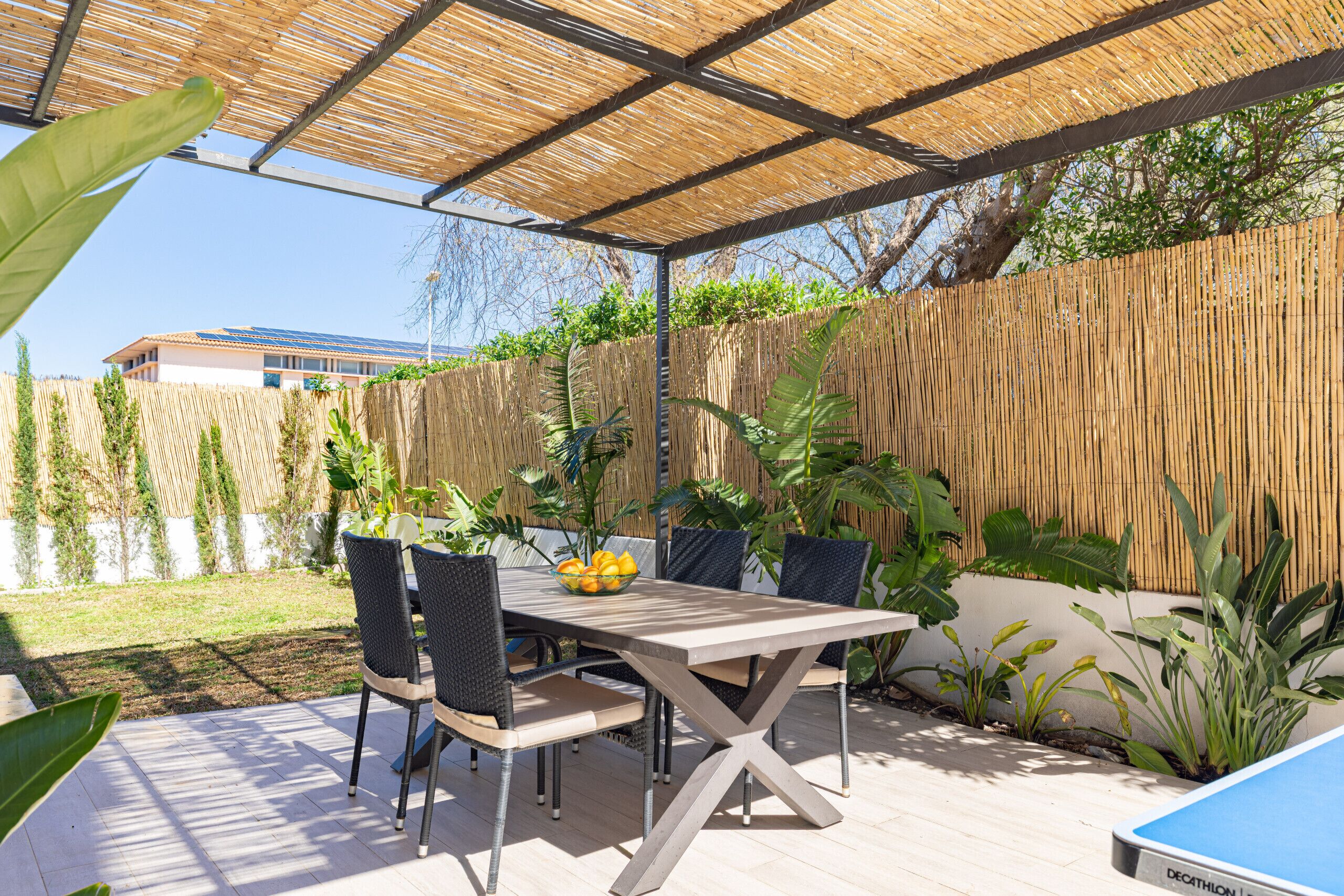 An outdoor patio with a table and chairs, surrounded by plants and a bamboo fence.