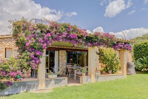 Una casa con fachada de piedra, un porche cubierto con mobiliario exterior y coloridas plantas florecientes de color púrpura.