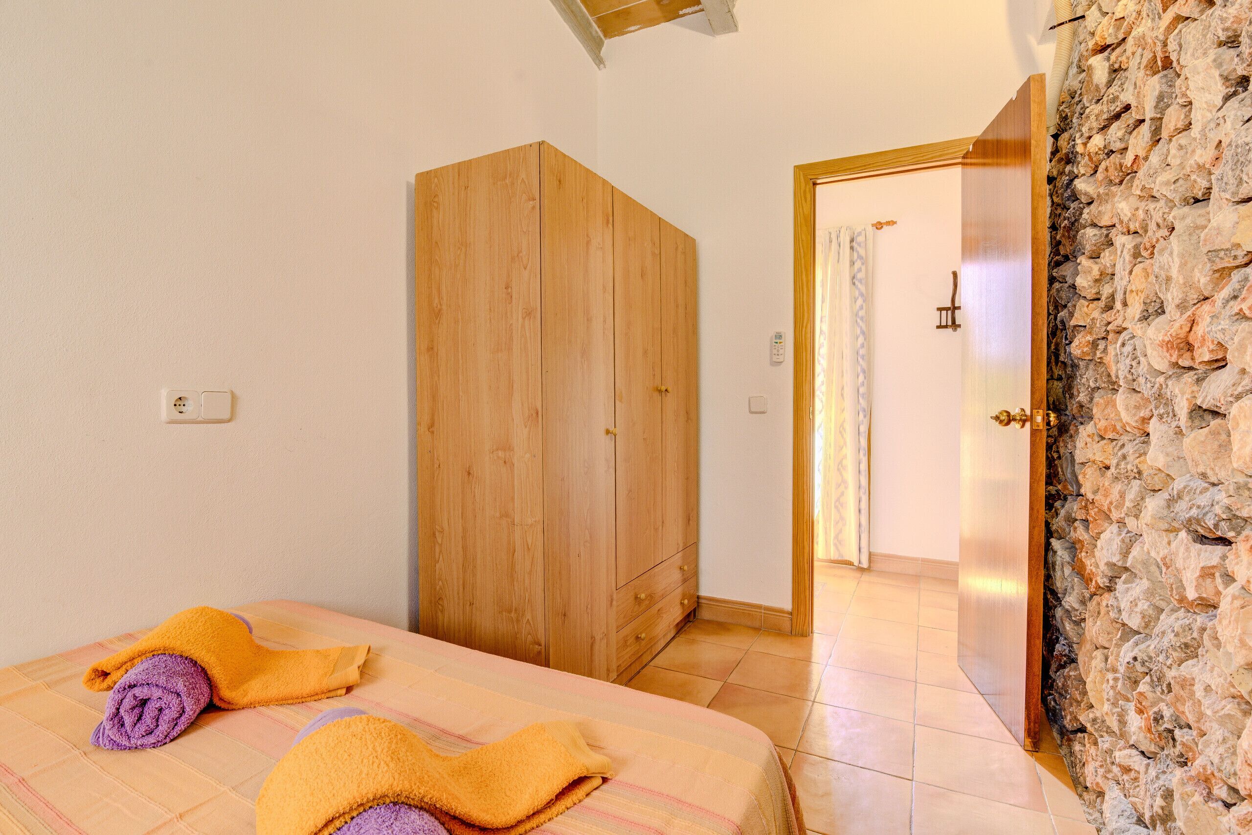 A room with a bed, wooden wardrobe, stone wall, and a door leading to another room.