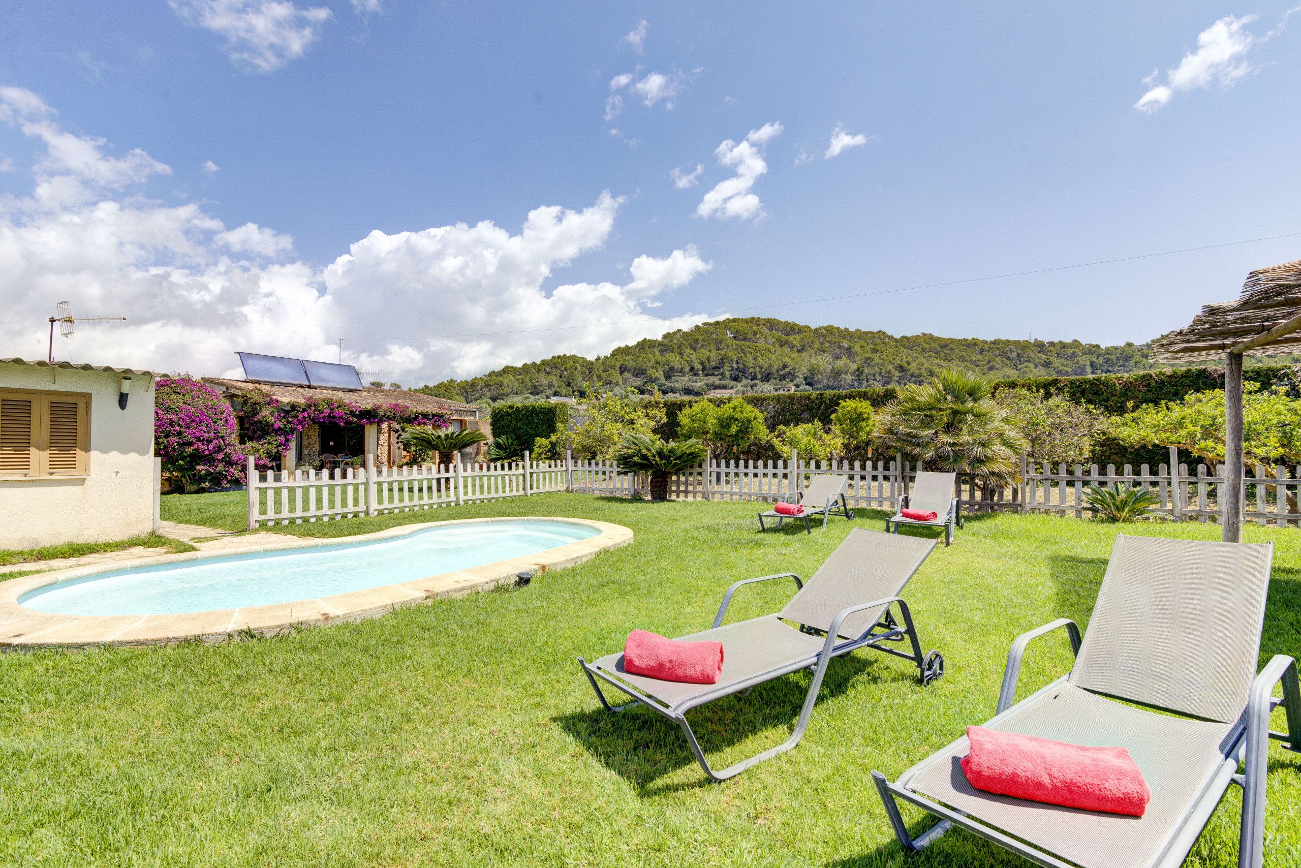 A backyard with a swimming pool, two lounge chairs with red cushions, a white picket fence, and a hill with trees in the background.
