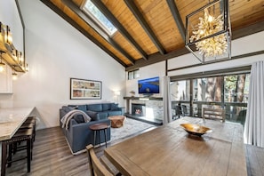 A modern living room with a fireplace, a dining area, and a large window overlooking trees.