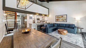 Ein geräumiger Wohnbereich mit einem Holzessttisch, einem blauen Ecksofa und einer Küche im Hintergrund.
