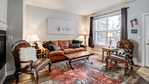 A living room with a fireplace, a sofa, a coffee table, two armchairs, a rug, and a large window with curtains. - Kingdom Park Townhome: Nice Space, On Shuttle Route (Breckenridge)