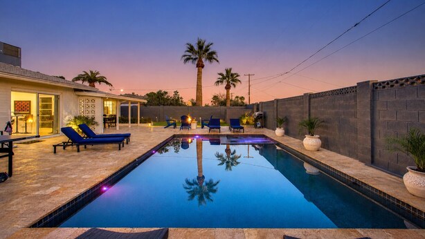 Una piscina con sillones de descanso, un porche cubierto y vistas a palmeras al atardecer.