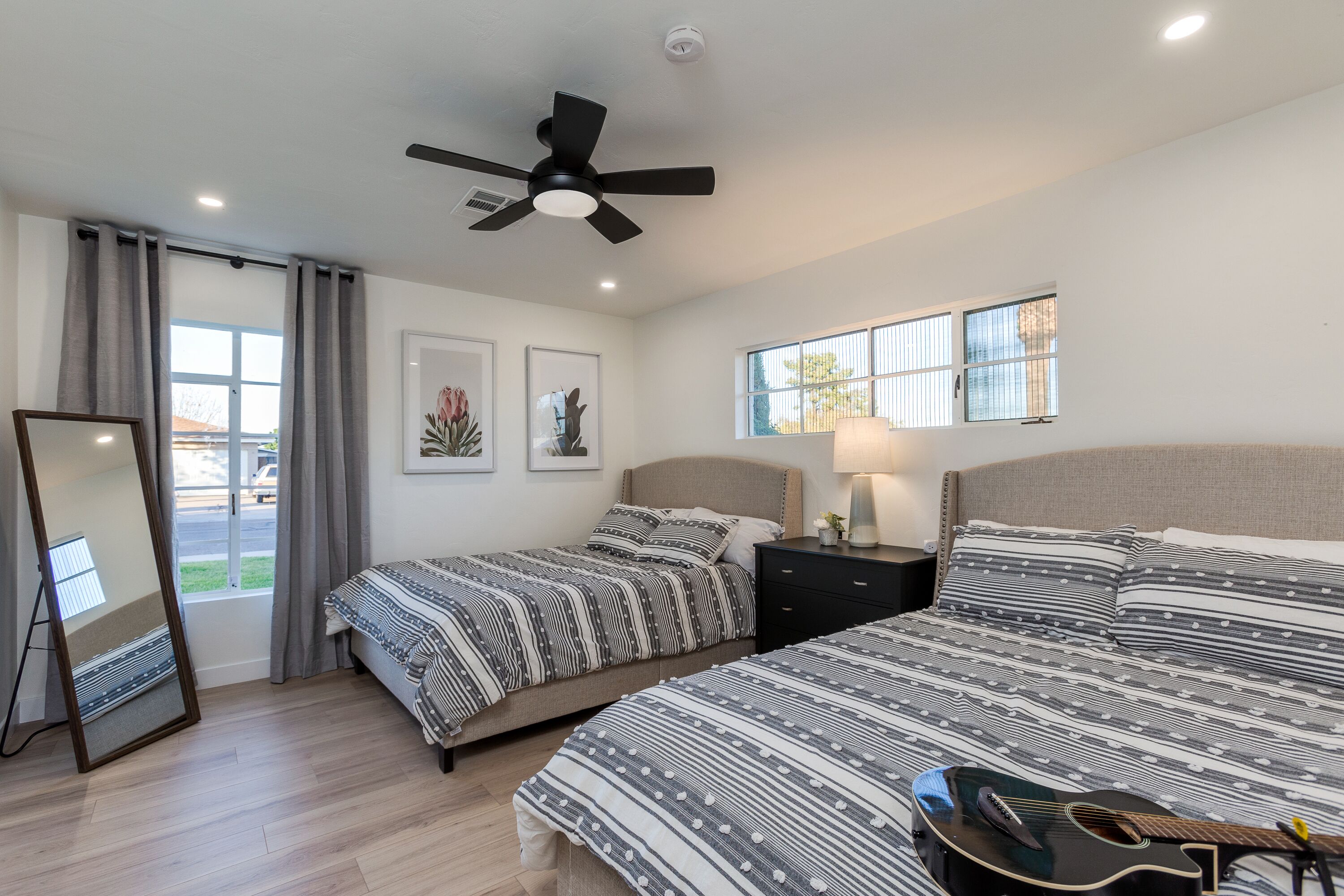 A bedroom with two beds, a ceiling fan, a window with blinds, a mirror, and a guitar on the bed.