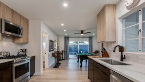 Una cocina moderna con estufa integrada, microondas y lavavajillas. Se observa una mesa de billar al fondo y un comedor con sillas.