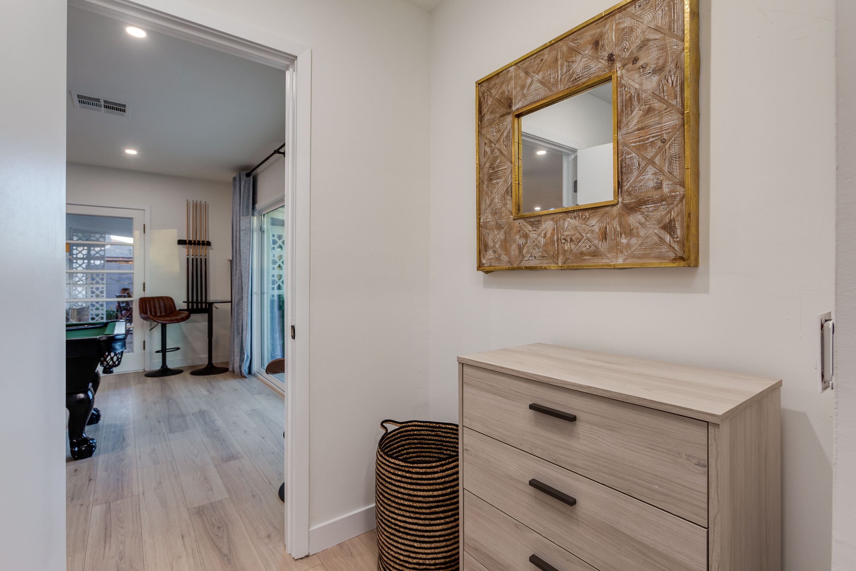 A modern room with a light wooden dresser, a mirror with a geometric frame, and a view into another room with a pool table.