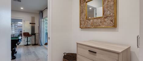 A modern room with a light wooden dresser, a mirror with a geometric frame, and a view into another room with a pool table.