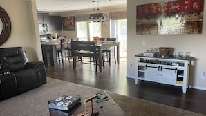 Un salon avec une cheminée, un coin repas avec une table et des chaises, une cuisine équipée de meubles et d’appareils électroménagers, et un grand tableau accroché au mur.