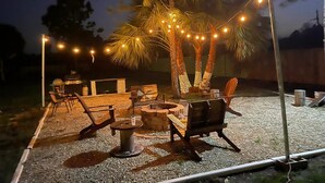 Un espace extérieur aménagé avec des chaises et un foyer, entouré de palmiers et illuminé de guirlandes lumineuses.