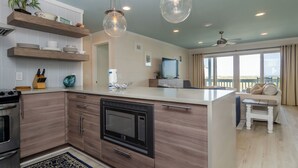 Een moderne keuken met houten kastjes, een inbouwoven en een magnetron. Er zijn hanglampen, een groot raam en een balkon met uitzicht.