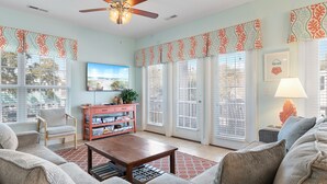A living room with a ceiling fan, a TV, a coffee table, and a bookshelf. - Walk to Beach 6BR Home w/ Private Pool & Beachside Porches (Surfside Beach)